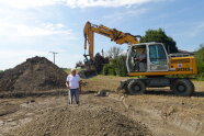 Ein Mann (der Bürgermeister) auf einer Baustelle, dahinter ein Bagger, in dem ebenfalls ein Mann sitzt.