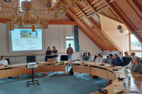 Gruppenpräsentation in einem großen Raum mit Holzdecke.